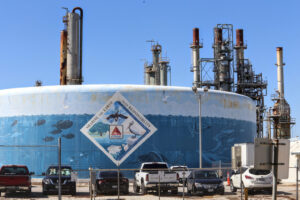A mural is seen painted on the side of a structure nearby smoke stacks at a refinery. The mural features fish, sea turtles, whales and shore birds in an ocean setting. A diamond-shaped section in the middle reads "sharing the earth with responsibility" and features the Citgo logo in the middle.