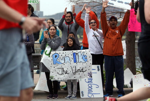 A photo of people standing on a sidewalk, cheering and holding a sign reading "run like the wind" as runners go by in front of them.