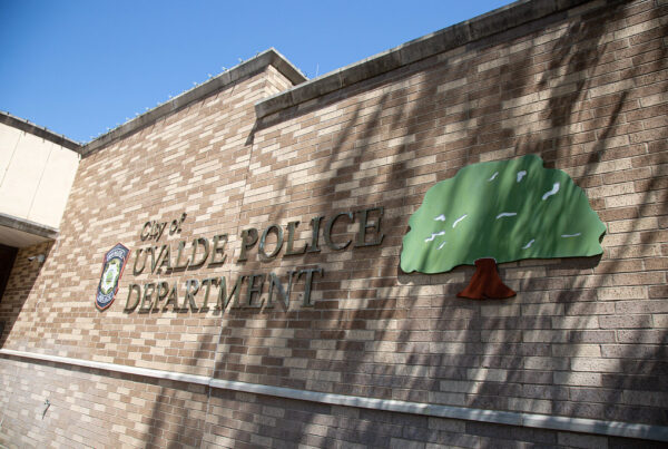 An exterior brick wall with the words "City of Uvalde Police Department." To the left of the words Is a police shield, and to the right is a tree.