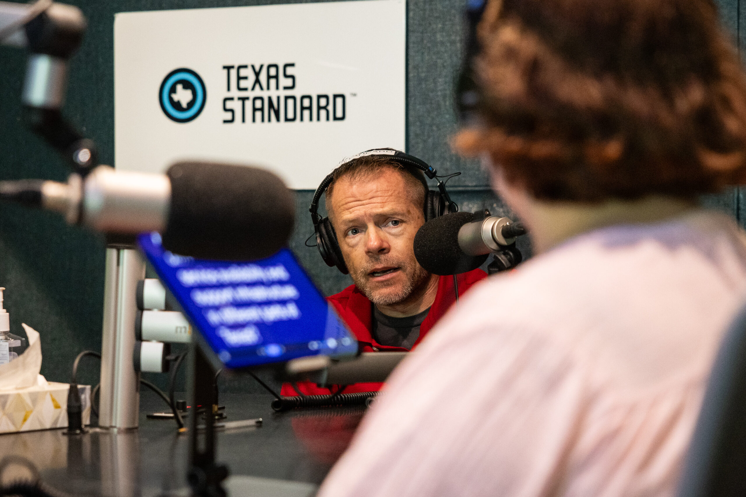A man wearing over the ear headphones speaks into a microphone looking in the direction of a woman seated facing him in the foreground. He is Chase Bearden, of the Coalition of Texans with Disabilities, and he is speaking to Texas Standard reporter and producer Shelly Brisbin during a disability advocates roundtable. They are in a radio station recording studio. The Texas Standard logo is seen on the wall behind Bearden and a tablet showing a teleprompter app is seen sitting on the table between him and Brisbin.
