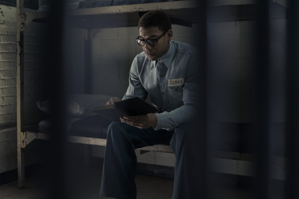 A photo of an actor playing Malcom X. The photo is taken through jail bars and it shows a man wearing glasses and reading a book in a denim prison uniform.