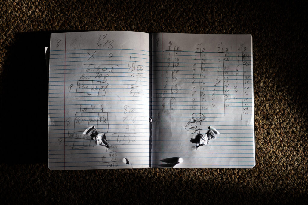 A close-up photo of an open notebook with numbers written in pencil and bullet holes tearing through the pages.