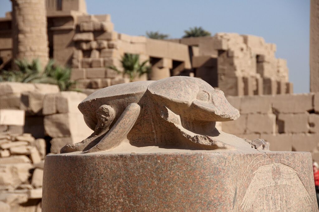 A close-up photo of a granite scarab beetle statue on a pedestal. Behind it are the bricks of a temple.
