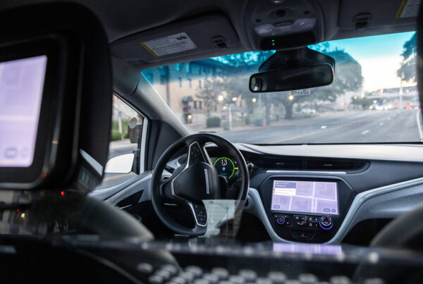 A photo from the backseat of a car shows a moving car with no one at the steering wheel. Two navigation screens are seen: one in the front console and another on the back of the headrest of the empty driver's seat.