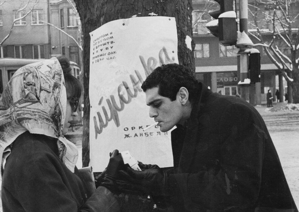 A black and white photo of a woman helping a man light a cigarette.