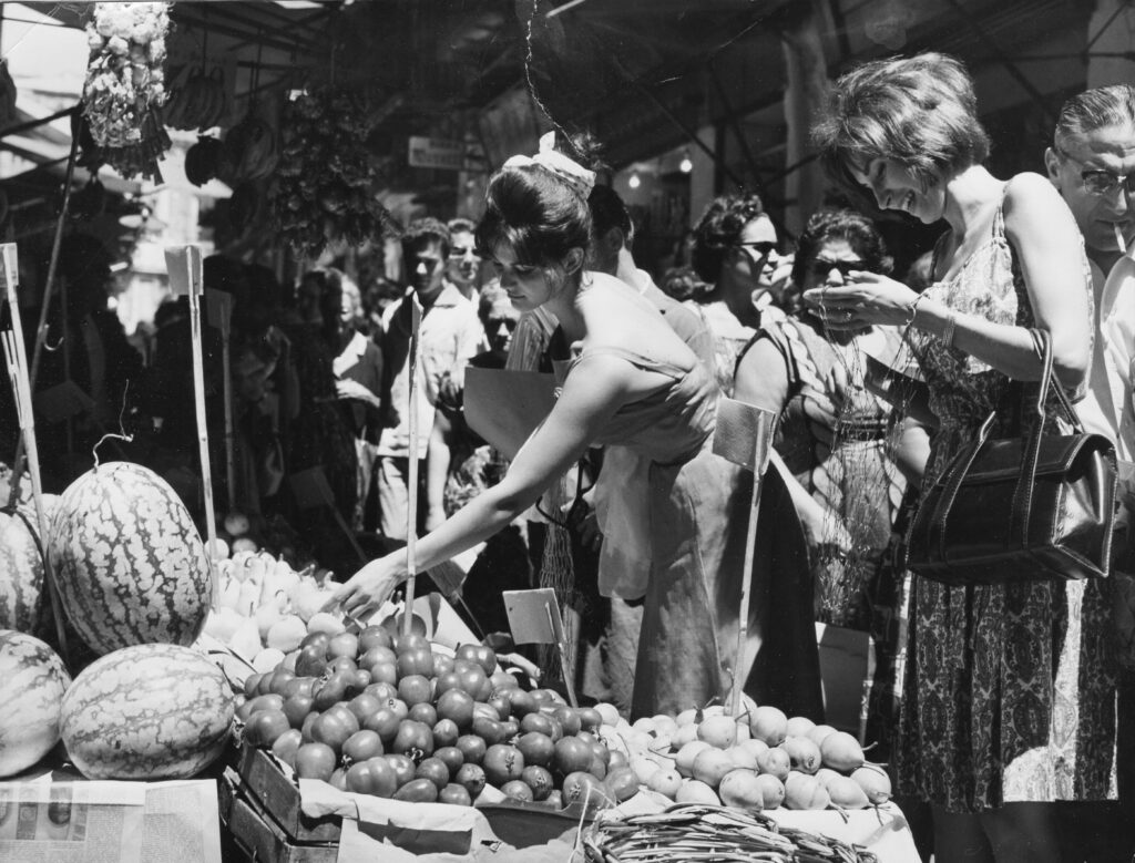 A black and white photo of two smiling women looking at produce in a busy, outdoor market.
