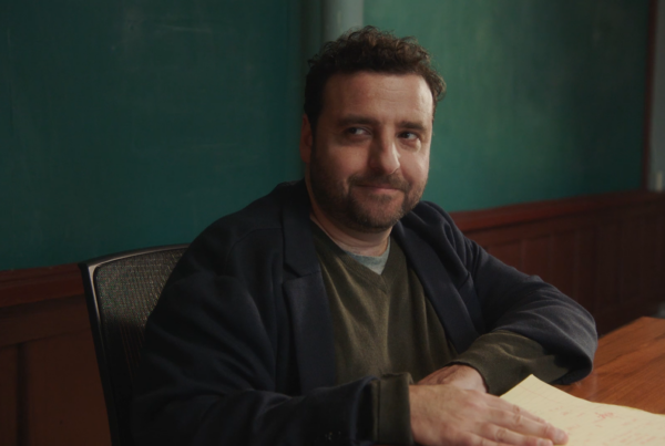 An actor sits at a desk with some paper. A green blackboard is behind him.