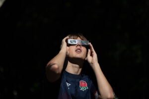 A man wearing eclipse glasses holds his hands up to his face as he looks up at the sky