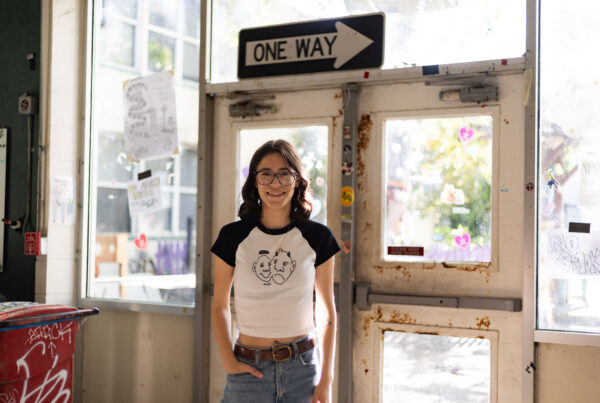 A portrait of a young woman standing in front of a door, below a "one way" sign. She has medium-length brown hair and glasses and is wearing a black-and-white T-shirt with two faces on it and jeans.