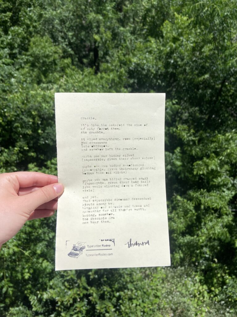 A photo of the typewritten poem on a torn half sheet of light yellow paper. A person is holding the paper up before a background of greenery.