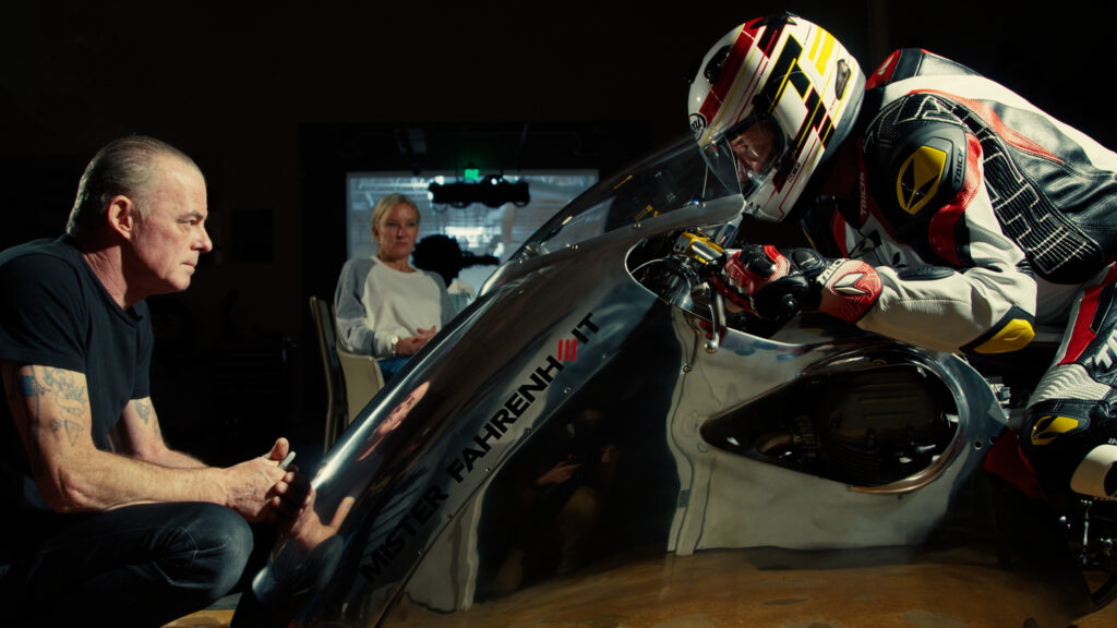 A man in full motorcycle leather and wearing a helmet sits atop a chrome motorcycle. In front of him is the motorcycle's designer and builder. And in the background is Haas's partner, Stacey Mayfield.