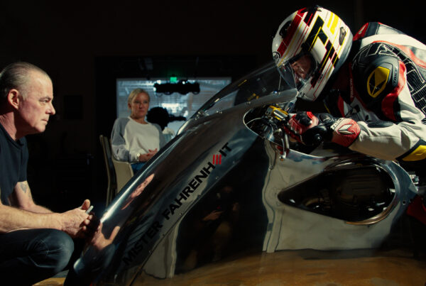 A man in full motorcycle leather and wearing a helmet sits atop a chrome motorcycle. In front of him is the motorcycle's designer and builder. And in the background is Haas's partner, Stacey Mayfield.