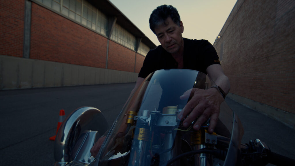 A man in a black t-shirt sits on a chrome motorcycle with a sidecar. He puts his hand on the windshield of the motorcycle. The sun sets in the background. He's in an alleyway between two long brick warehouse-type buildings.