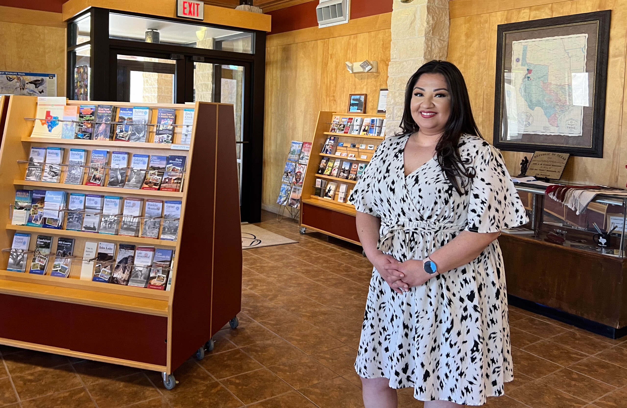 A woman poses indoors for a photo. To the left of her are shelves lined with travel brochures.
