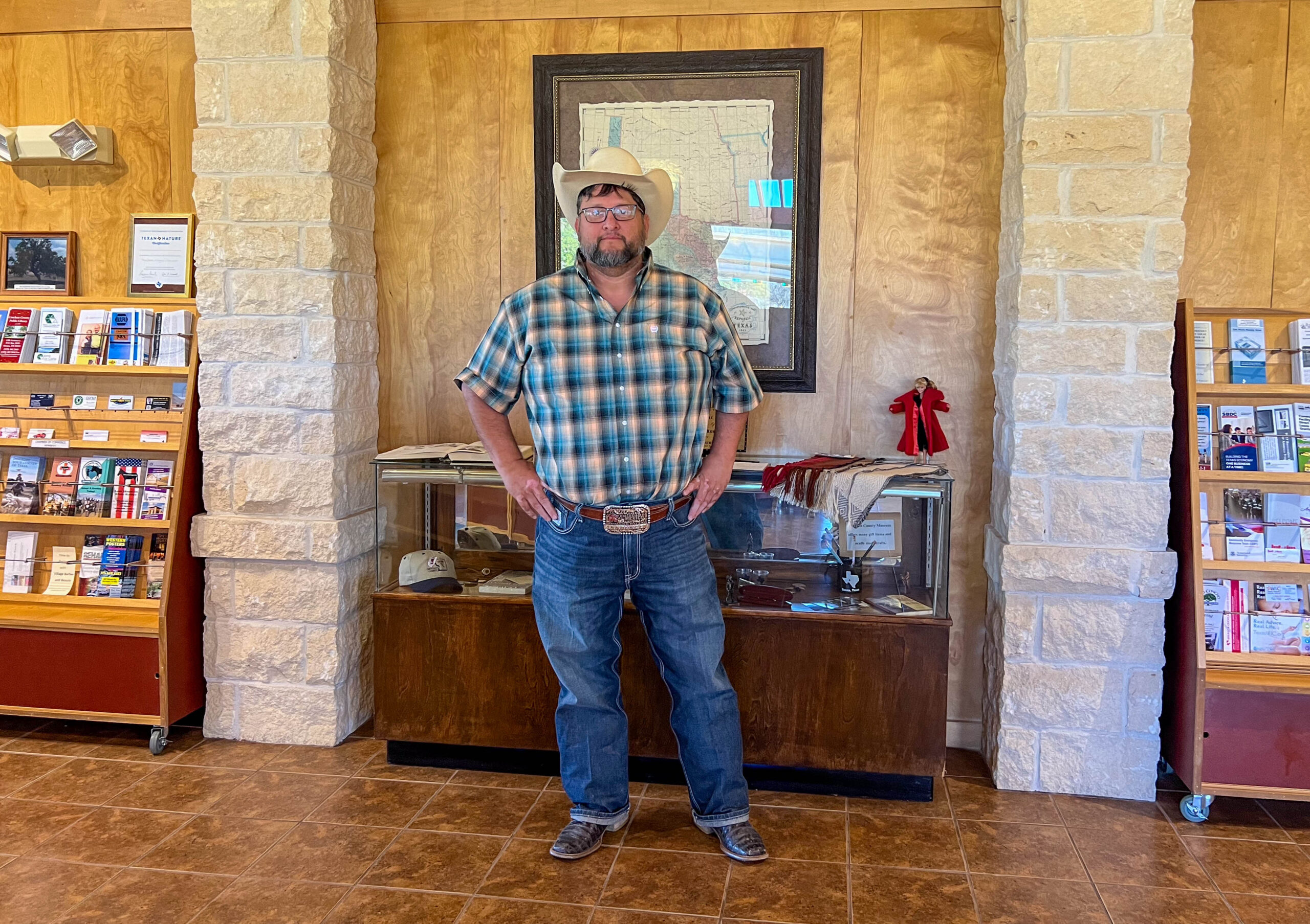 A man poses indoor for a photo. He's wearing several traditional Texas wear, including a cowboy hat, large belt buckle, and cowboy boots. This is Ruben Cruz.