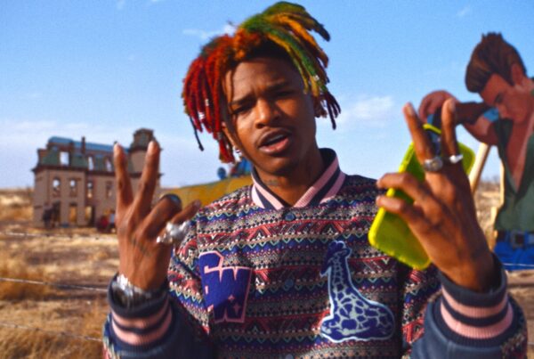 A photo of a young man with colorful locs in his hair posing for the camera in a colorful jacket before the background of the field where the movie "Giant" was filmed.