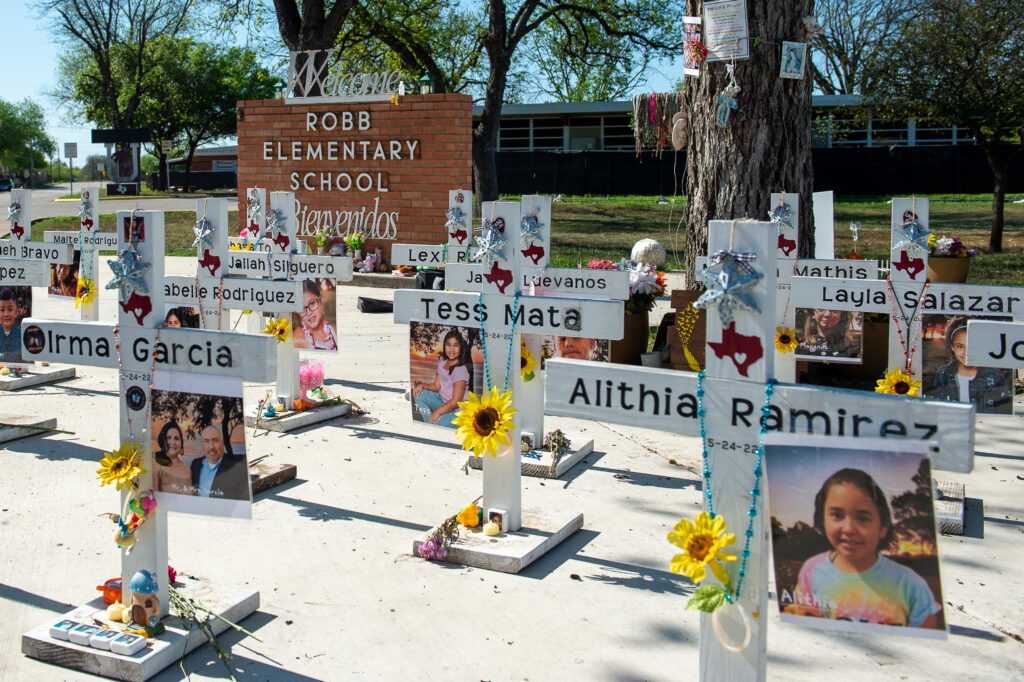 A photo of white crosses with photos and flowers in front of Robb Elementary School.