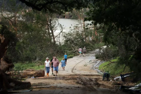People walk on a road covered with mud and fallen tree branches near a swollen river.