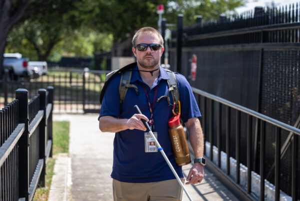 A man wearing sunglasses and holding a cane walks towards the camera on a sidewalk.