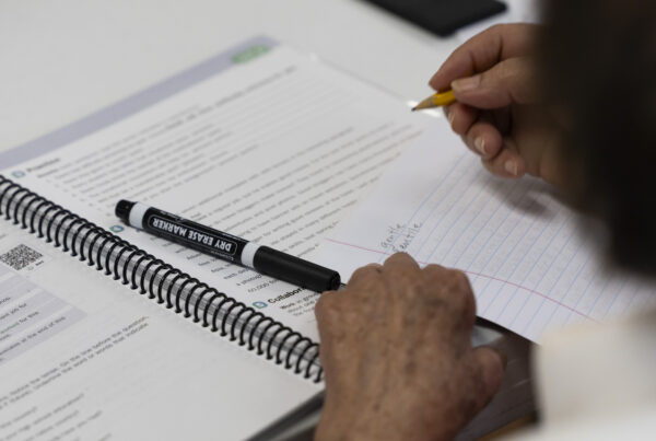 Hands are seen writing in pencil on lined paper on top of a spiral workbook