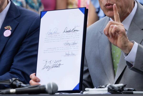A closeup of a document with several signatures on it, held up by a man whose other hand is pointing up.