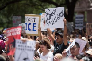 A man surrounded by a group of fellow protesters holds up a sign that says "No human is illegal!!"