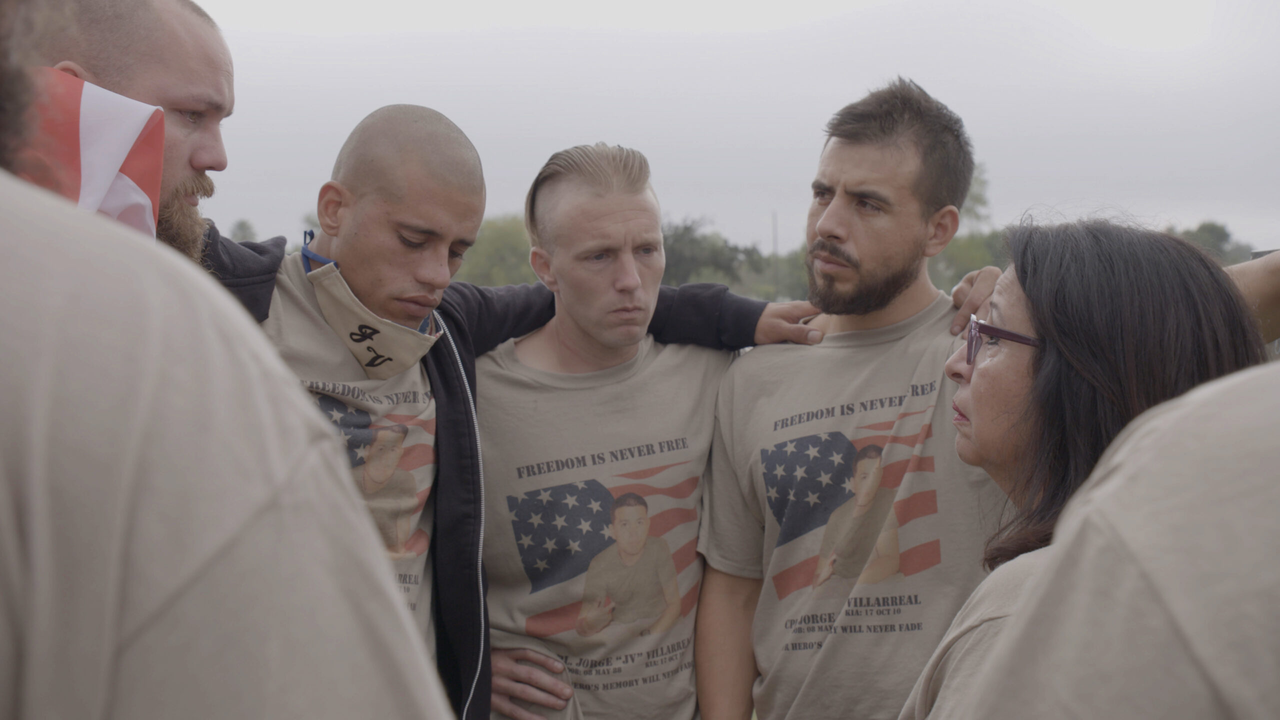 A photo of people gathered in a tight circle. They are wearing matching shirts with an American flag and a photo of their fellow Marine, JV Villarreal.