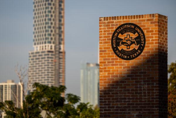 The sun shines on a brick wall with a Huston-Tillotson University emblem on it as city buildings are illuminated in the background.