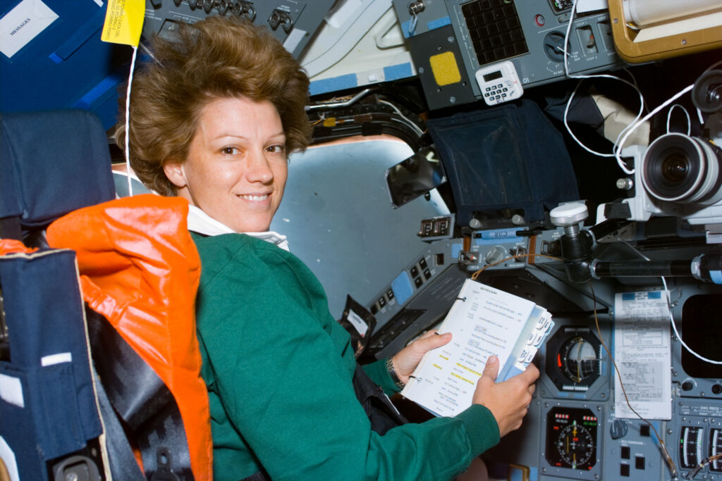 Astronaut Eileen Collins, mission commander, looks over a procedures checklist at the commander's station on the forward flight deck of the Space Shuttle Columbia on Flight Day 1. She smiles at the camera and her hair stands on end as she floats in space.