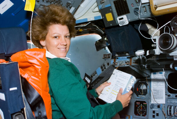 Astronaut Eileen Collins, mission commander, looks over a procedures checklist at the commander's station on the forward flight deck of the Space Shuttle Columbia on Flight Day 1. She smiles at the camera and her hair stands on end as she floats in space.