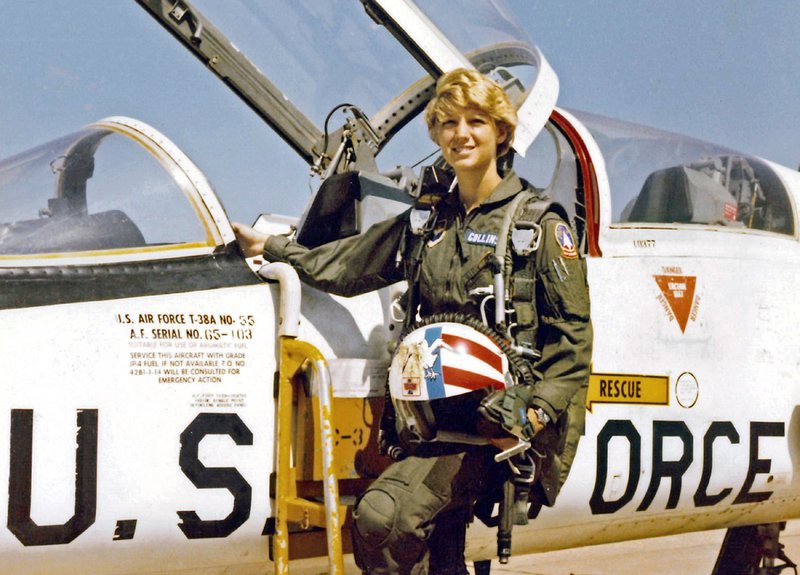 Collins wears a flight suit and holds her helmet as she stands next to the open cockpit of her plane.