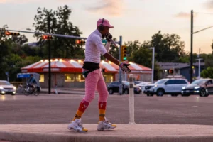 A man wearing bright athletic clothes, wearing headphones and holding a microphone is at a street corner as the sun sets.