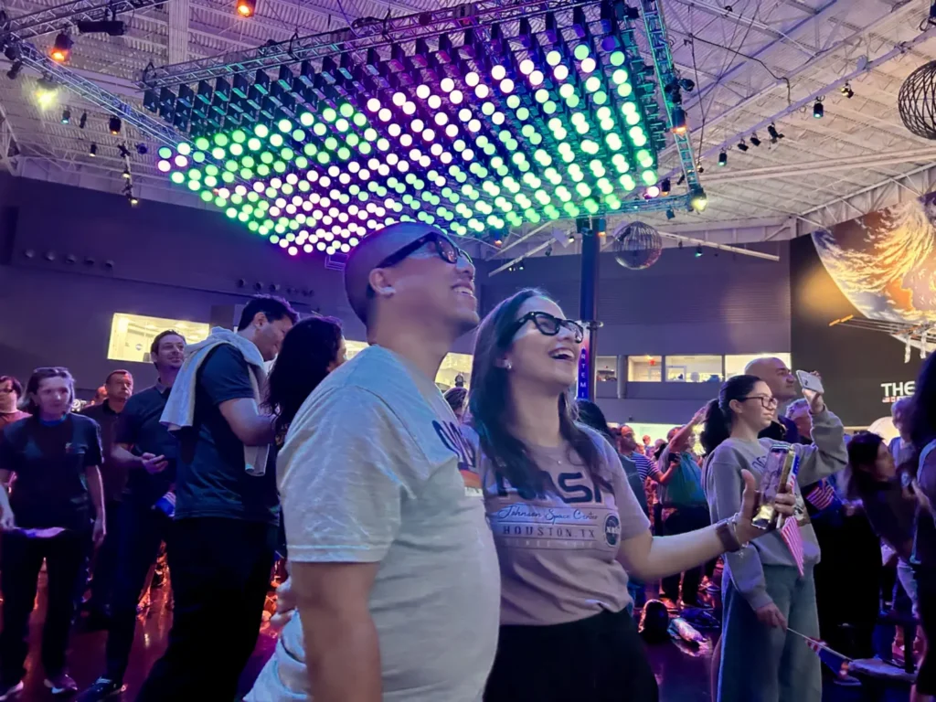 Two people wearing NASA shirts are front and center in a crowd watching a screen in awe. The room is lit with colorful lights.