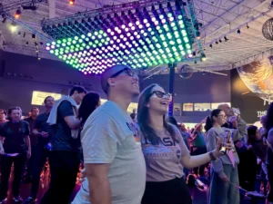 Two people wearing NASA shirts are front and center in a crowd watching a screen in awe. The room is lit with colorful lights.