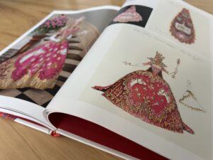 A close-up photo of an open book shows the sketch of an elaborate pink gown on one page and a photo of a woman in the same gown on the other page.