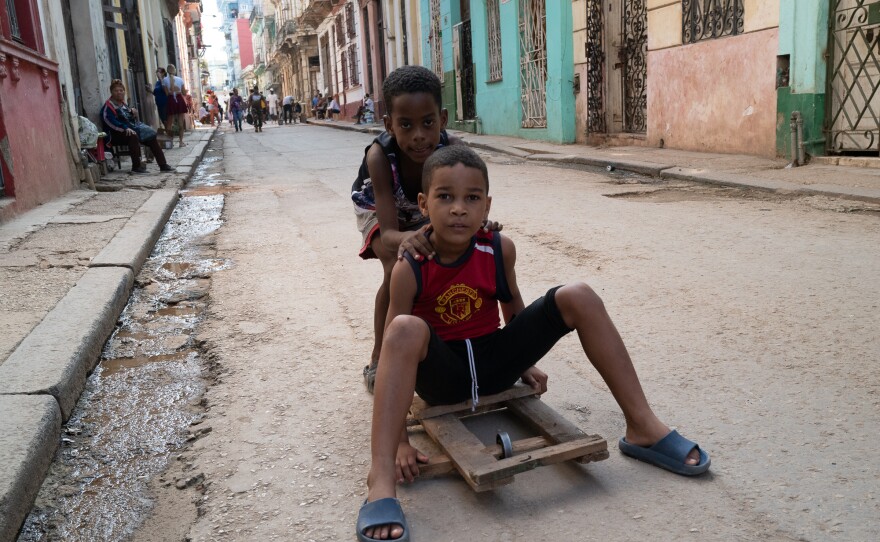 Two kids ride on a makeshift skateboard past colorful buildings on a rough-textured street in Havana..