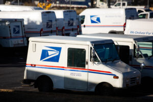 Shadows fall on a fleet of white mail carrier trucks and vans in a parking lot.