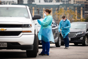 cars line up at a covid 19 testing site in austin, with two health care workers wearing blue gowns and face masks helping patients