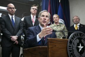 a white man with graying hair wearing a blue suit--the texas governor- sits in a wheelchair at a lectern with the texas round state seal on it with four members of texas government behind him
