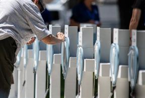 An angled photo shows several white wooden crosses with a light blue heart and white wooden design on the front. Each cross has a name on it and a Sharpie hanging from it. A person is bending over slightly to write on one cross.