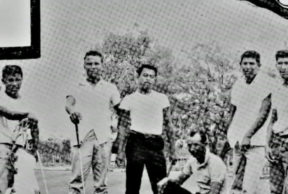 A grainy black and white photo shows five young mean in t-shirts and jeans posing with golf clubs. A man who may be their coach, kneels in the center.
