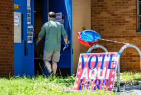 A voter walks into the blue double doors of a school building past a "Vote Aqui Here" sign and two red and blue star-shaped balloons.